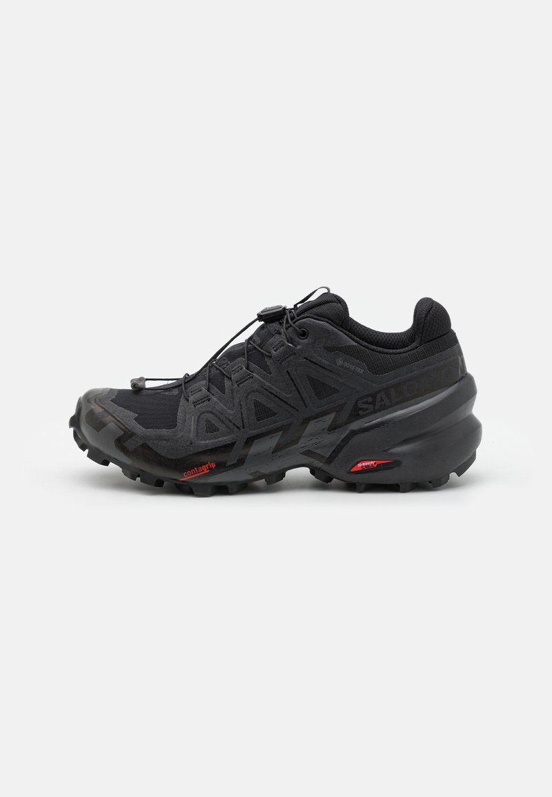 Chaussure de trail noire avec semelle robuste, tige en mesh, superpositions géométriques, lacets à serrage rapide et discrètes touches de rouge sur fond blanc.