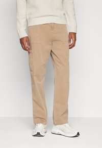 Beige Cargo-Hosen mit seitlichen Taschen, aus weichem Baumwollstoff. Lockere Passform, gerades Bein-Design, kombiniert mit weißen Sportschuhen.