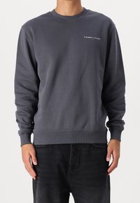 Homme portant un sweat à manches longues gris foncé avec le logo "TOMMY JEANS" sur le haut de la poitrine gauche et un jean noir, se tenant devant un fond uni.