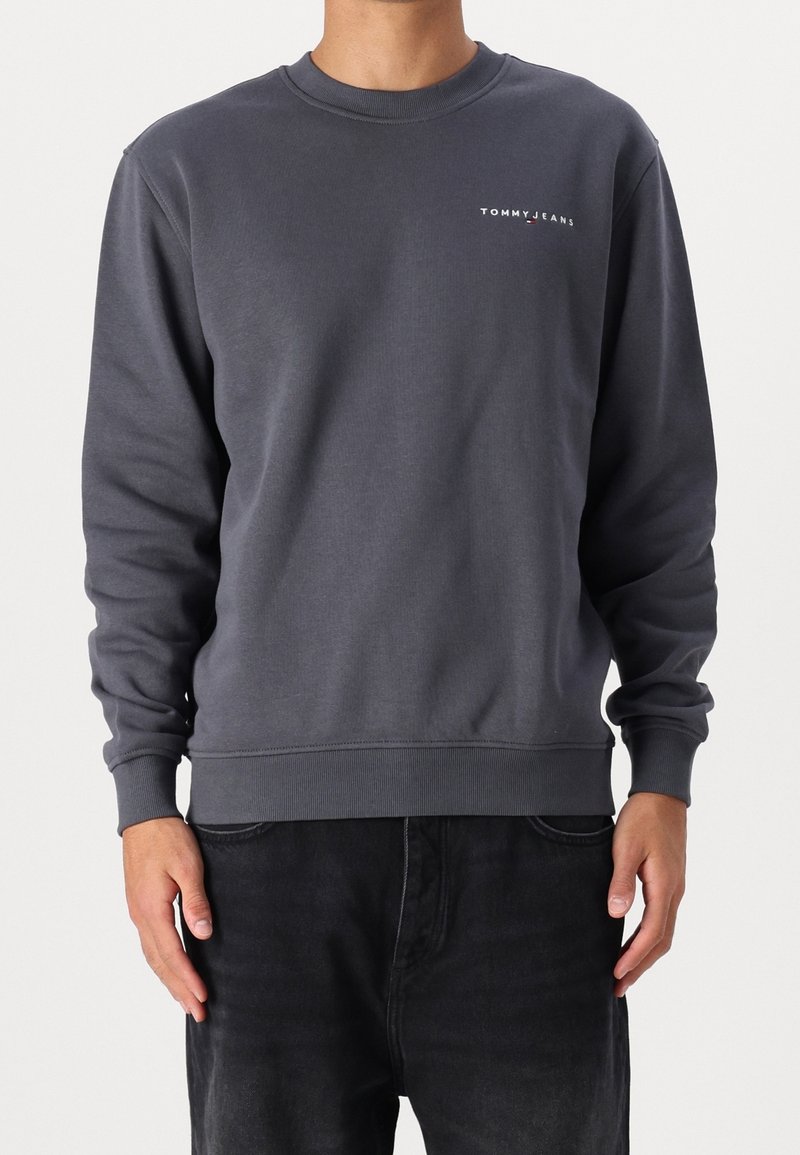 Homme portant un sweat à manches longues gris foncé avec le logo "TOMMY JEANS" sur le haut de la poitrine gauche et un jean noir, se tenant devant un fond uni.