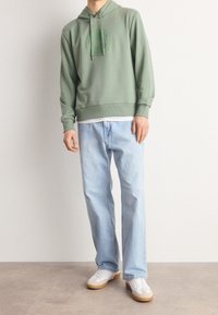 Person som bär en ljusgrön hoodie med dragsko, vit undertröja, lösa ljusblå jeans och vita sneakers med gummisulor som står på ett grått golv.