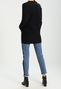 Musta pitkähihainen pusero teksturoidusta kankaasta, pyöreällä kaula-aukolla ja napilla takana. Yhdistettysin sinisiin farkkuihin ja nilkkureihin.