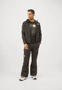 Črno jakno na zadrgo preko črne grafične majice z rumenim motivom sonca, kombinirano z ohlapnimi črnimi hlačami in belimi supergami.