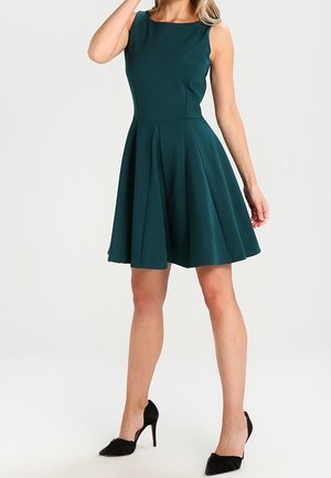 Ärmelloses, dunkelgrünes knielanges Kleid mit plissiertem Rock, getragen mit schwarzen High-Heels an einer stehenden Frau.