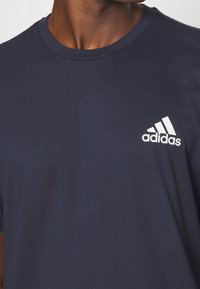 Námorní modré bavlněné tričko s kulatým výstřihem a krátkými rukávy. Na levé části hrudi se nachází bílá značka Adidas. Hladká textura.