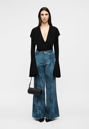 Žena v čiernom top s hlbokým výstrihom do V a dlhými rozšírenými rukávmi, vysokým pásom široké modré džínsy, čierne špicaté topánky a s malou čiernou kabelkou v ruke.