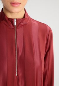 Burgundowa koszula na zamek błyskawiczny z stójką, o gładkiej satynowej fakturze i pionowych paskach na materiale. Detal z zamkiem podkreśla przód.