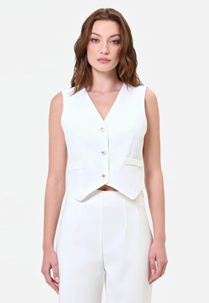 Giovane donna con capelli castani mossi che indossa un gilet bianco senza maniche con bottoni e pantaloni bianchi a vita alta abbinati, in piedi su sfondo bianco.
