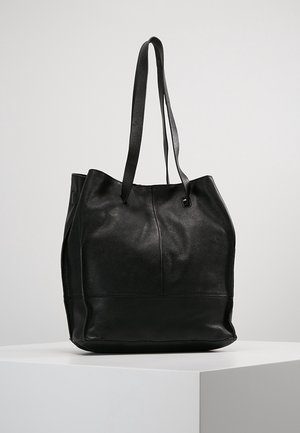 Černá kožená taška přes rameno s dlouhými popruhy, vystavená na bílém podstavci na světlém jednobarevném pozadí.