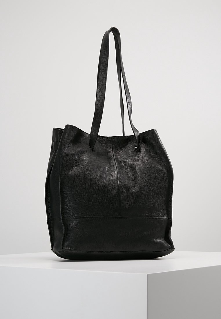 Sac fourre-tout en cuir noir avec de longues anses, présenté sur un socle blanc contre un fond clair uni.