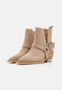 Botas de tobillo de ante marrón claro con punta afilada, una correa con hebilla y un tacón bajo apilado. Presentan un detalle metálico en el tacón. Textura suave.