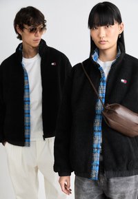 Black fleece jacket with a zippered front and ribbed cuffs, layered over a blue and black checkered shirt. Brown crossbody bag included.