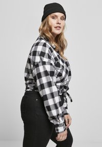 Mustriga mustvalge flanellpits, millel on seotud vöökoht, koos mustade teksadega. Mudel kannab musta ribaga beanie mütsi.