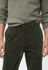 Pantalones de pana verde con canalones verticales, cierre de botones y bolsillos frontales. Combinados con un suéter de punto gris claro.