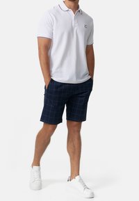 Man in een wit poloshirt, marineblauwe geruite shorts en witte sneakers, staand met de handen in de broekzakken op een witte achtergrond.