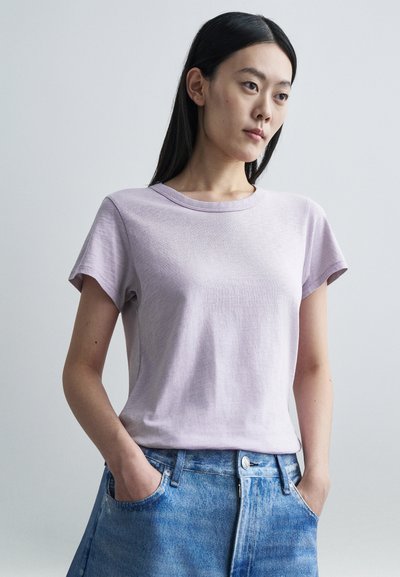 Femme aux cheveux longs et foncés portant un t-shirt violet clair et un jean bleu, debout avec les mains dans les poches devant un fond clair uni.