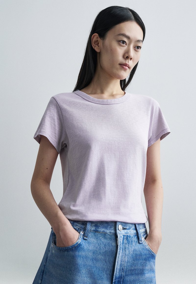 Mulher de cabelos longos e escuros, vestindo uma t-shirt lilás clara e jeans azuis, em pé com as mãos nos bolsos contra um fundo claro simples.