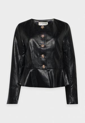 Le modèle pose devant un fond clair et uni, portant une veste en cuir noire à basque avec des boutons dorés, un pantalon à jambes larges et des chaussures noires.