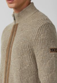 Beige gebreide trui met een hoge kraag, voorzien van een textuurpatroon en een contrasterende bruine streep aan de voorkant. Leren logo patch op de mouw.