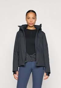 Giubbotto isolato nero con cappuccio, dotato di zip frontale e polsini elasticizzati. Indossato sopra una maglia nera e un pantalone blu, dalla texture liscia.