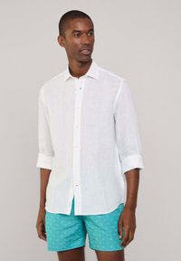 Camicia bianca di lino a maniche lunghe con colletto button-down, maniche arrotolate e bottoni beige chiaro. Indossata con pantaloni corti turchesi a fantasia.