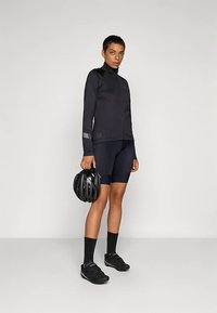 Svart cykeljacka med långa ärmar, kroppsnära design, texturerade detaljer, bärs med svarta cykelshorts, svarta strumpor och håller en hjälm.