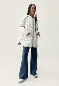 Prešívaná bunda v světle krémové barvě, s kapucí, předním zipem a dvěma bočními kapsami. Kombinovaná s širokými modrými kalhotami a bílými teniskami.