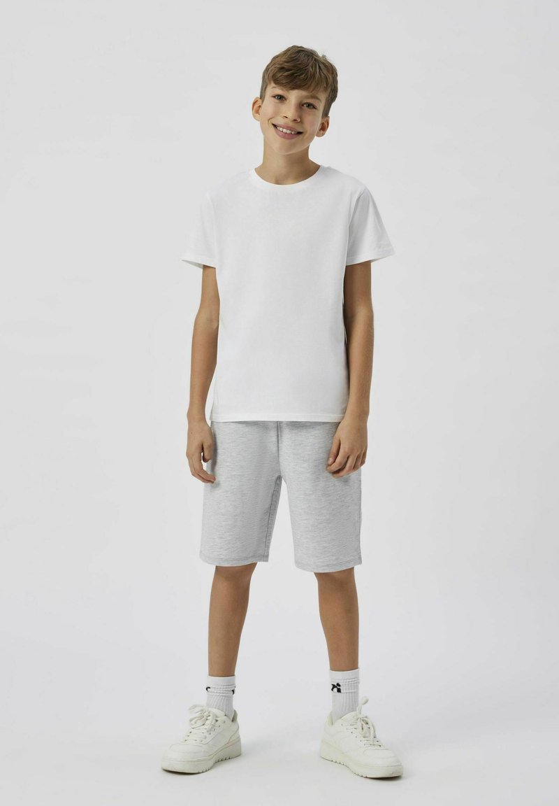 Vit bomulls-t-shirt, gråa träningsshorts och vita sneakers på ett barn som står mot en neutral bakgrund.