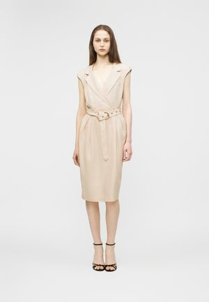 Kvinde med langt brunt hår iført en ærmeløs beige knælængde kjole med bælte og sorte sko med stropper mod en ensartet lys baggrund.