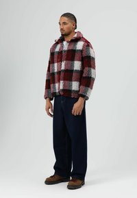 Rood-zwart-wit geruit fleecejack met kraag, knopen aan de voorkant en een gezellige textuur. Draagt bij donkere denim broek en bruine laarzen.