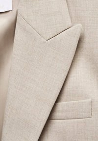 Dettaglio di un blazer in lino beige con revers a punta, tessuto strutturato e una discreta tasca sul petto. Linee pulite con un aspetto sartoriale.