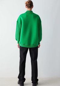 Suéter de punto verde de gran tamaño con mangas texturizadas, cuello y dobladillo acanalados, combinado con pantalones negros y zapatos negros gruesos. Vista trasera.