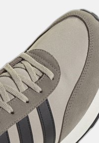 Baskets beiges avec des superpositions en daim, rayures noires sur le côté et lacets beiges, montrant les détails de l'avant et du côté sur fond blanc.