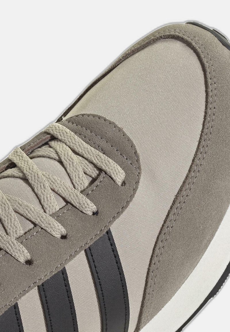 Beige sneaker met suède overlays, zwarte strepen aan de zijkant en beige veters, met zowel voor- als zijdetails op een witte achtergrond.