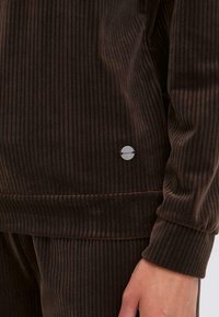 Bruine corduroy trainingsjack met ribbelstructuur, voorzien van een ronde zilveren logo-accent en een ronde hals. Glad, zacht materiaal.