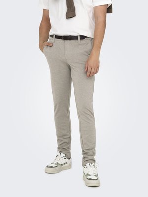 Bărbat purtând pantaloni bej slim fit, pantofi sport albi, cămașă albă cu mâneci scurte și un pulover maro așezat pe umeri, stând cu o mână în buzunar.