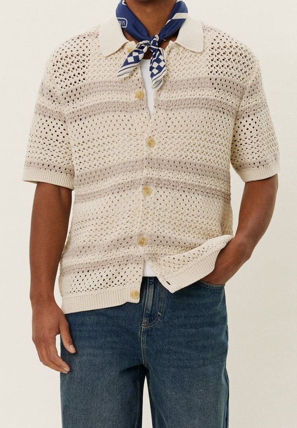 Homme portant une chemise beige en maille ajourée à manches courtes avec boutons, un jean bleu et un foulard bleu et blanc à motifs autour du cou.