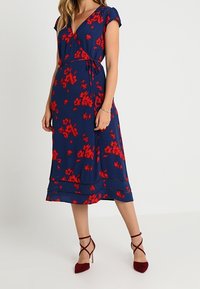 Kvinna klädd i en marinblå omslagsklänning med röd blommönster, korta ärmar och vinröda högklackade ankelremsskor.