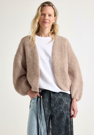 Kvinde iført en beige strikket cardigan, hvid skjorte og blå jeans med sort blomstret blonder-overflade, stående med den ene hånd i lommen.