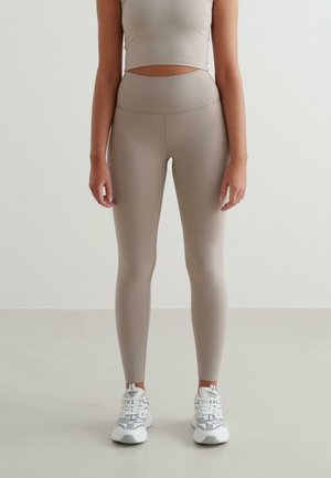 Beige naadloze sportoutfitset bestaande uit een cropped top en high-waisted leggings. Gladde textuur met subtiele branding. Model draagt witte sneakers.