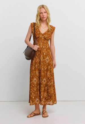 Vrouw met lang blond haar, gekleed in een bruine, met patroon mouwloze maxi-jurk, bruine crossbodytas en sandalen, staand tegen een effen achtergrond.