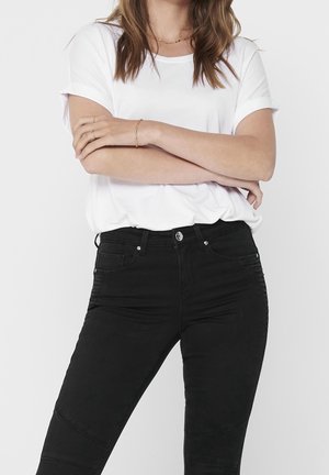 Kvinde iført en hvid kortærmet skjorte og sort højtaljede jeans med armene krydset foran en ensfarvet baggrund.