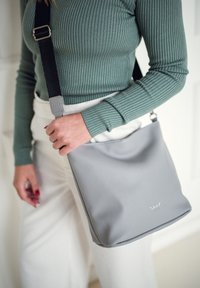 Person in einem gerippten grünen Langarmoberteil und weißen Hosen, hält eine graue Umhängetasche mit einem schwarzen Riemen über dem Oberkörper.