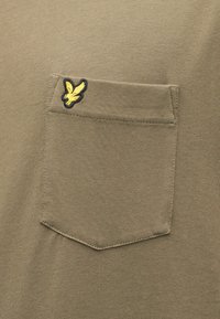 Olívazöld rövid ujjú ing mellzsebbel. A zsebén hímzett sárga logó látható. Puha, texturált anyagból készült, testhez simuló fazonban.