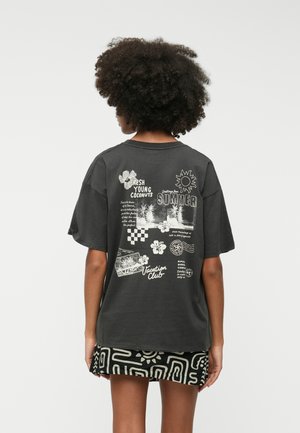 T-shirt surdimensionné gris foncé avec des impressions graphiques blanches présentant des thèmes tropicaux, y compris du texte, des fleurs et des motifs géométriques au dos.