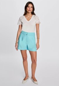 Weißes Oberteil mit Spitzenärmeln und V-Ausschnitt, kombiniert mit hellblauen, plissierten Shorts mit einem taillierten Bund. Komplett mit weißen Schuhen.