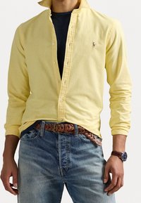 Man med en oknäppt gul skjorta över en svart t-shirt, blå jeans med brunt flätat bälte, och en brun klocka på höger hand.