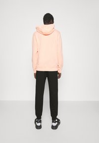 Světlá růžová mikina s kapucí na šňůrku, nošená s černými tepláky a černými sandály. Hladká látka, volný střih, minimalistický design.