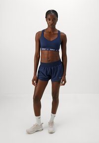 Donna atletica in piedi, con indosso un reggiseno sportivo blu navy e pantaloni corti, calze Nike bianche e scarpe Nike rosa chiaro su uno sfondo semplice.