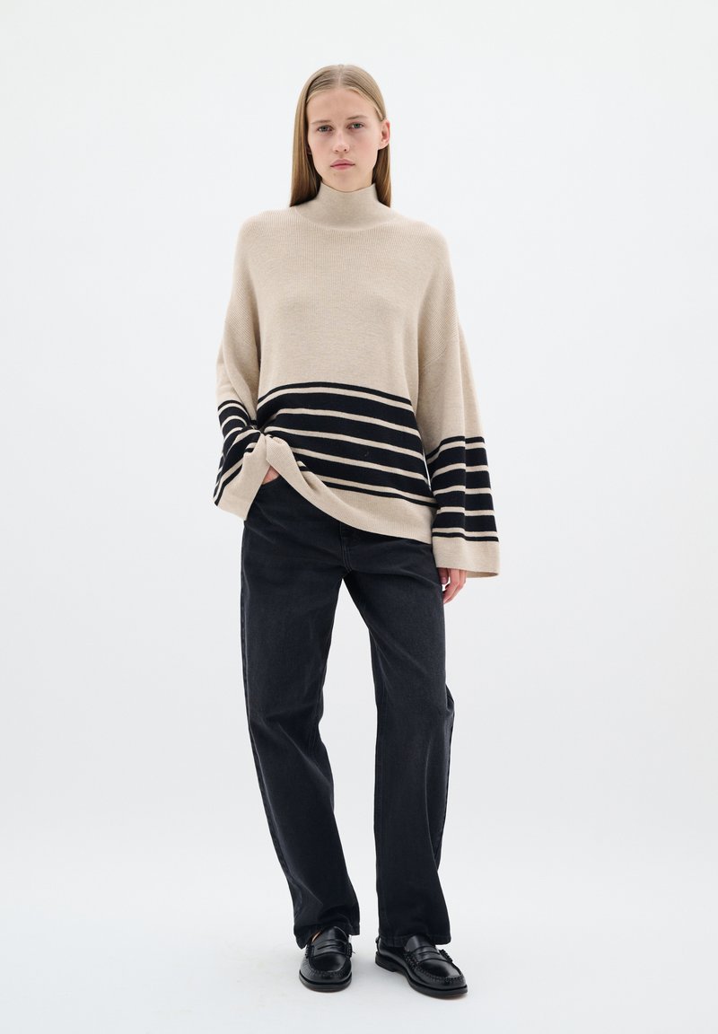 Beige gebreide trui met een hoge hals en zwarte horizontale strepen, gecombineerd met zwarte rechte jeans en zwarte schoenen.